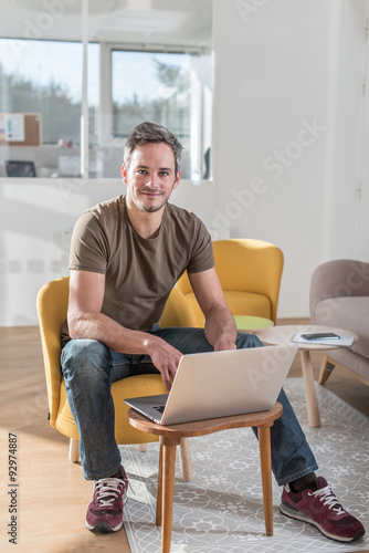 Portrait of a middle aged grey hair man with beard in a stylish vintage living room with wooden floor. He is sitting in an orange chair with his laptop in front of him He is wearing casual clothes