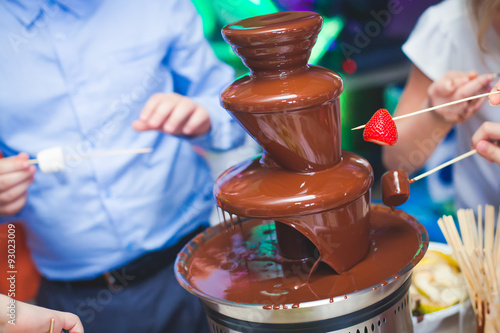 Vibrant Picture of Chocolate Fountain Fontain on childen kids birthday party with a kids playing around and marshmallows and fruits dip dipping into fountain 