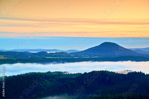 Blue foggy deep valley after rainy night. Rocky hill bellow view point. The fog is moving between hills and peaks of trees, blue clouds in the sky.