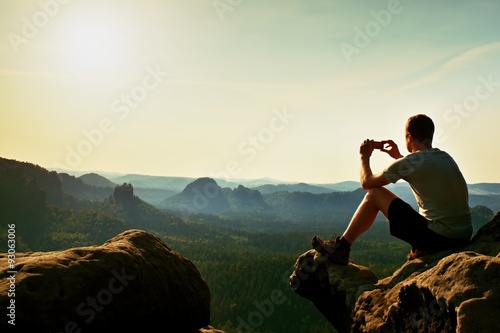 Tourist in grey t-shirt takes photos with smart phone on peak of rock. Dreamy hilly landscape below, orange pink misty sunrise in a beautiful valley below rocky mountains.