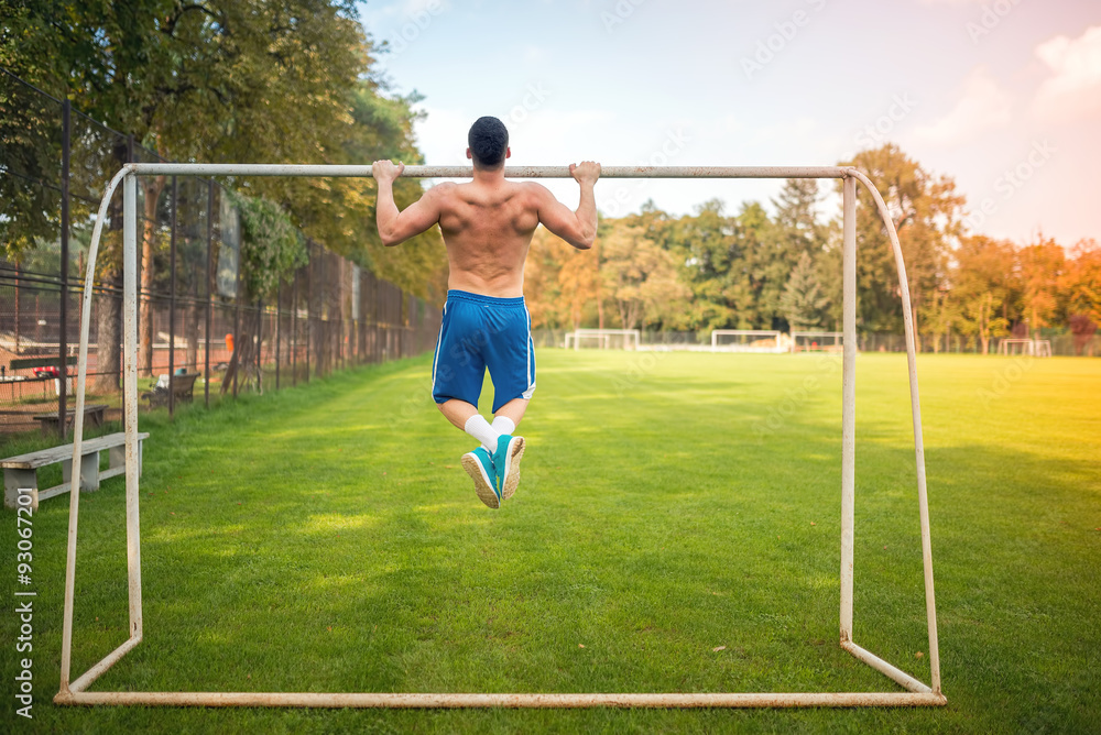 Young muscular man doing pull ups on football court, hardcore training outdoors