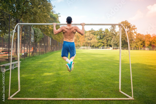 Young muscular man doing pull ups on football court, hardcore training outdoors