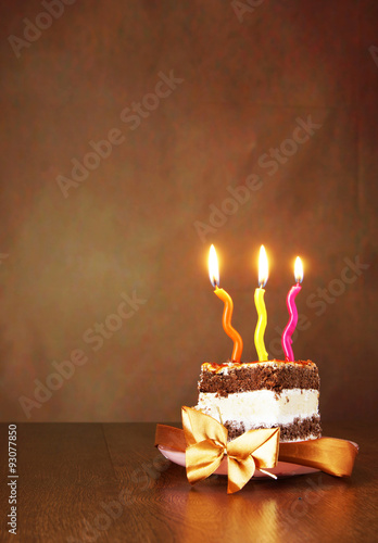 Piece of birthday chocolate cake with three burning candles against brown background