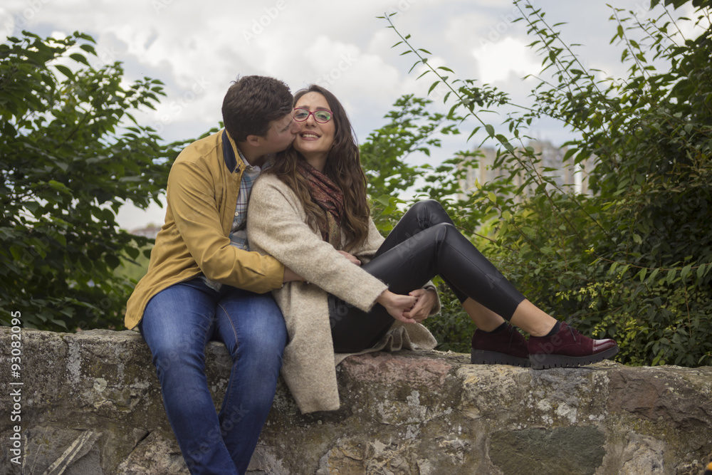 Young hipster couple sitting on stone wall, kissing and cuddling