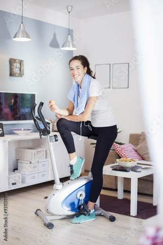 Sporty woman training on exercise bike in living room having an apple