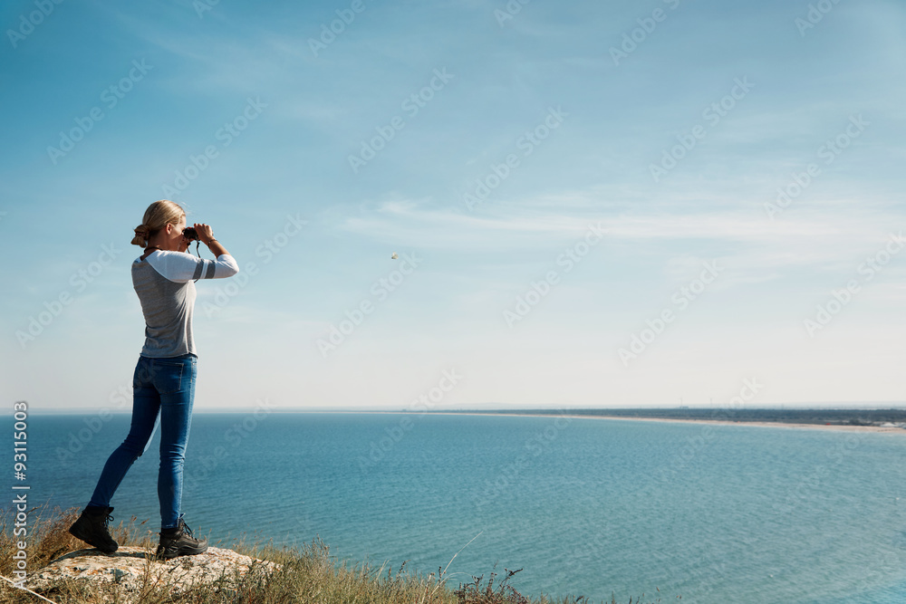 Girl with binoculars at the mountains in the rays of the evening sunset. Young beautiful woman on nature background. She is resting in the open air. Keep holding binoculars. Look ahead. Copy space
