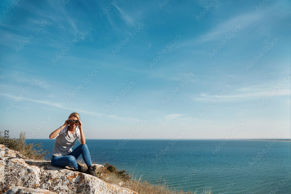 Young active female standing on the top of mountain and watching away in binocular, extreme lifestyle, traveling and trek concept, copy space