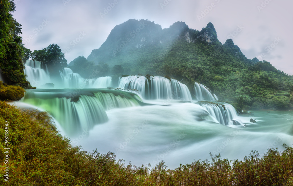 Fototapeta premium Ban Gioc Waterfall craggy limestone permissive side misty morning with foreground grass and tones of the lower cascade. It is considered the most beautiful waterfalls in Southeast Asia