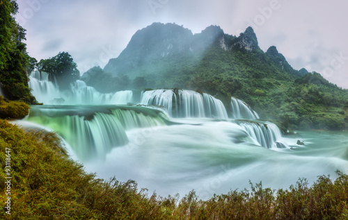 Ban Gioc Waterfall craggy limestone permissive side misty morning with foreground grass and tones of the lower cascade. It is considered the most beautiful waterfalls in Southeast Asia