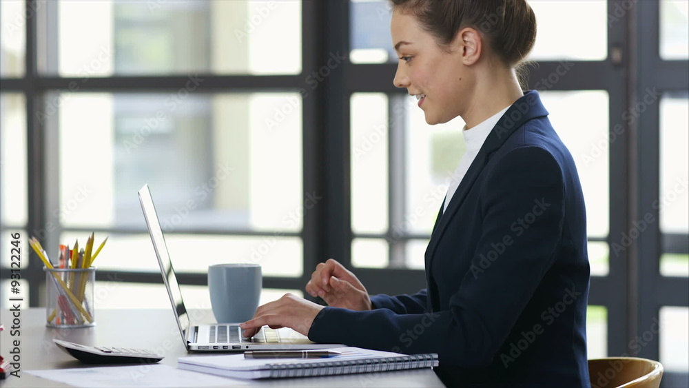 Tilt up slow motion shot of female professional using laptop. Side view of smiling businesswoman typing on laptop keyboard. She is sitting at desk in brightly lit office. Tilt up slow motion shot of female professional using laptop. Side view of smiling businesswoman typing on laptop keyboard. She is sitting at desk in brightly lit office.