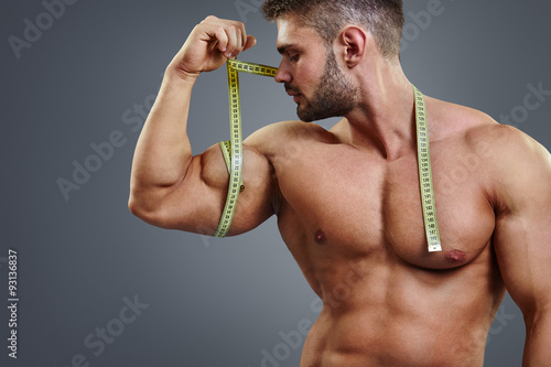 Closeup handsome strong athlete measuring muscle biceps with tape measure isolated over gray background. Bodybuilder tries to measure bicep by himself.