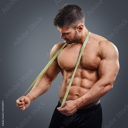 Muscular sports man tries to measure his neck muscles with measuring tape  isolated over gray background. Confused bodybuilder holding tape measure.  Stock Photo