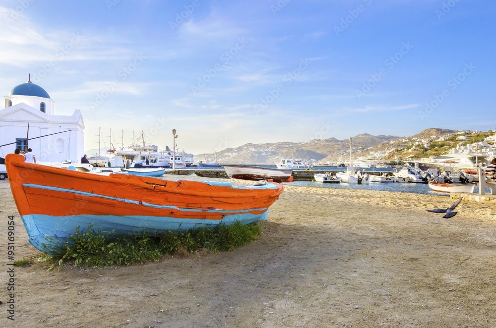 Fototapeta premium Colourful fishing and sail boats anchored on the Chora port in Mykonos,Greece. View of orange,blue boat on sand shore and the whitewashed blue dome church of Hora harbour, a greek island landscape.