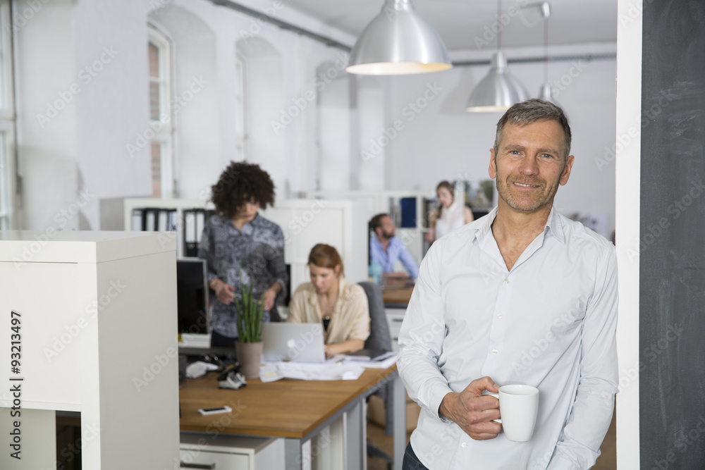 Smiling man in office with colleagues in background Smiling man in office with colleagues in background