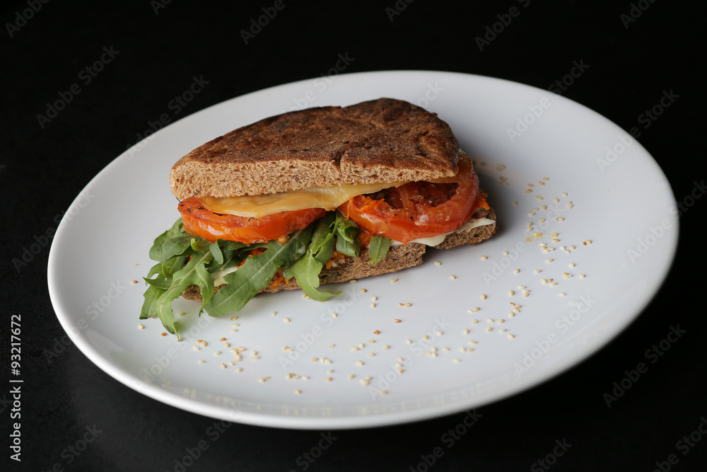 Vegetarian sandwich with tomato, arugula and cheese on white plate on dark background.