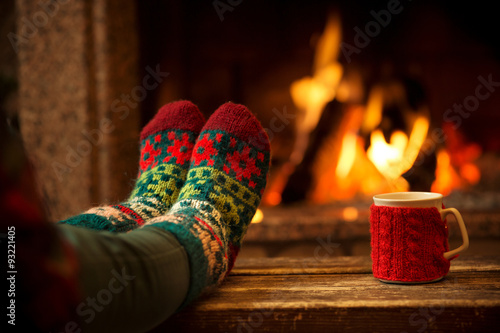 Feet in woollen socks by the Christmas fireplace. Woman relaxes by warm fire with a cup of hot drink and warming up her feet in woollen socks. Close up on feet. Winter and Christmas holidays concept. photo