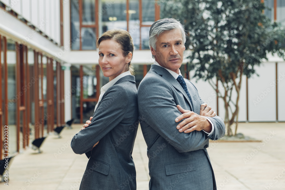 Mature business man and woman standing back to back looking at camera Mature business man and woman standing back to back looking at camera