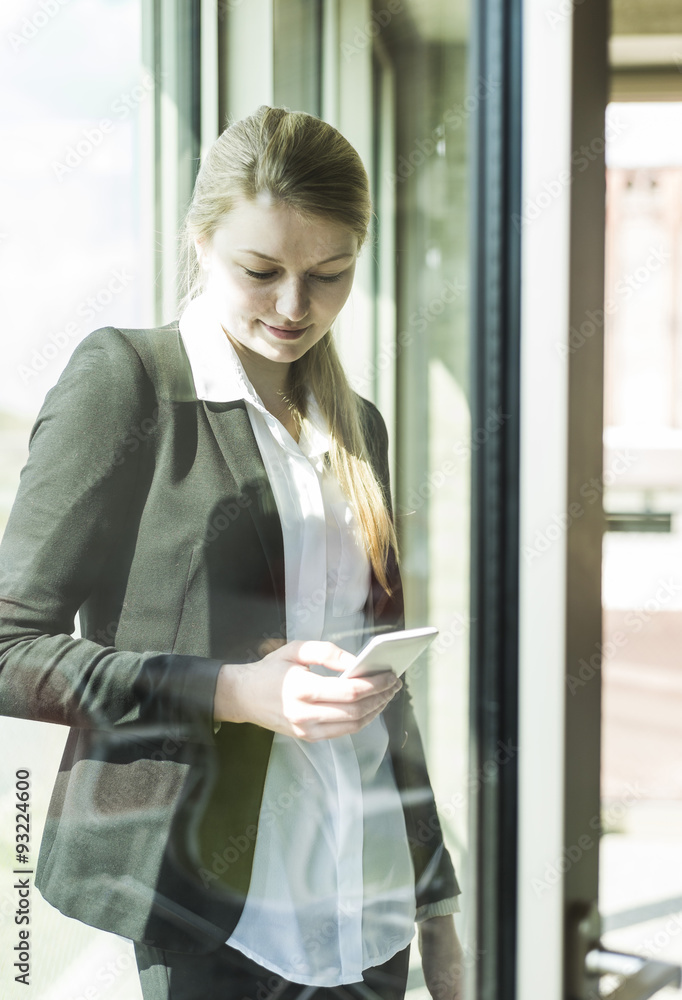 Smiling young businesswoman at the window looking on cell phone