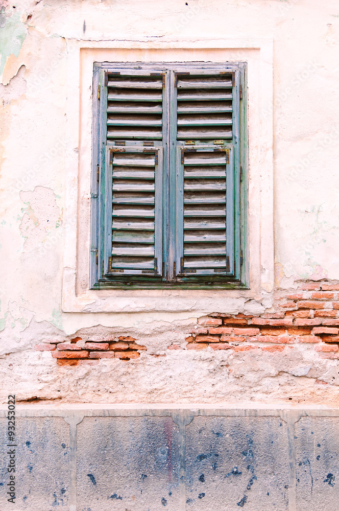 Old pale blue slatted window in a brick wall with the white washed rendering eroded in parts.