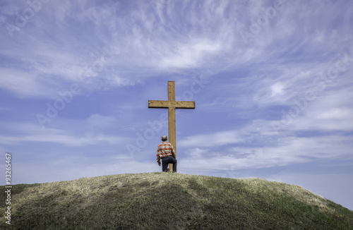 horizontal image of a man kneeling by a wooden cross on a grassy hill surrounded by a beautiful blue sky with wispy white clouds floating by in the summer time.