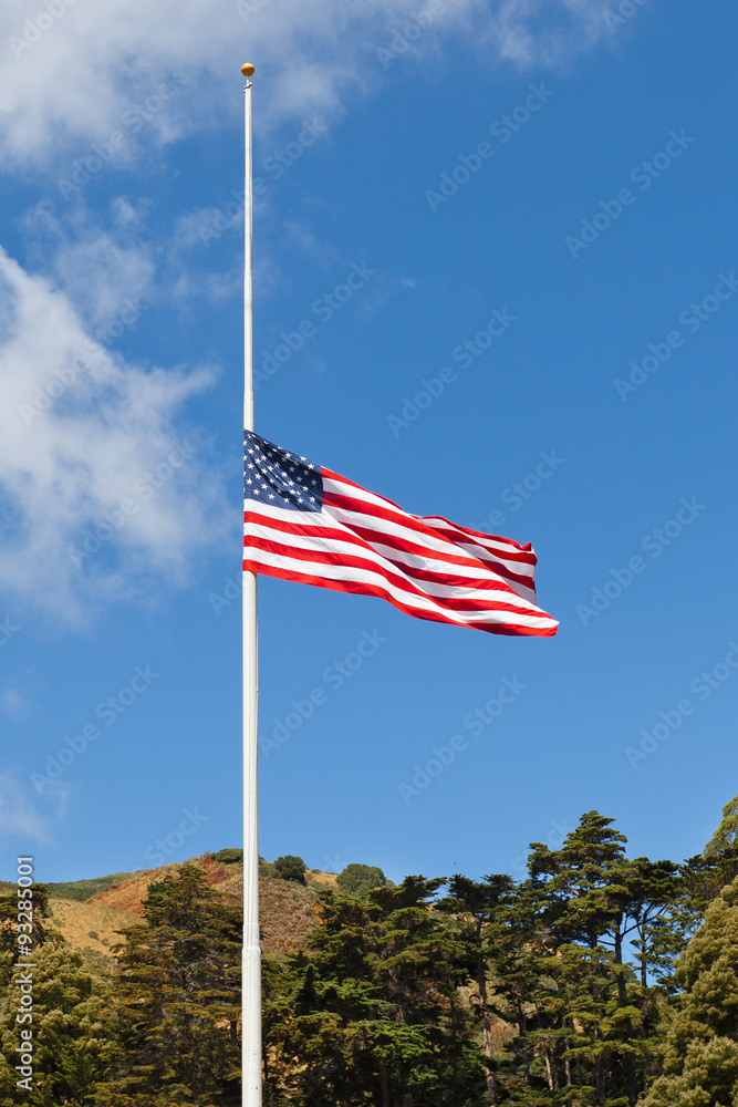 American flag flying at half mast against blue sky. Tree covered hills in the background.