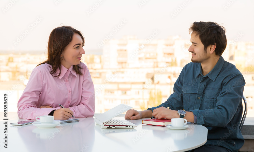 Fototapeta premium Informal Business Meeting at Roof Top Cafe Man and Woman Casual Clothing Sitting at White Round Table with Coffee Mugs Laptop Notepads and Telephone Discussing Smiling Making Hand Notes