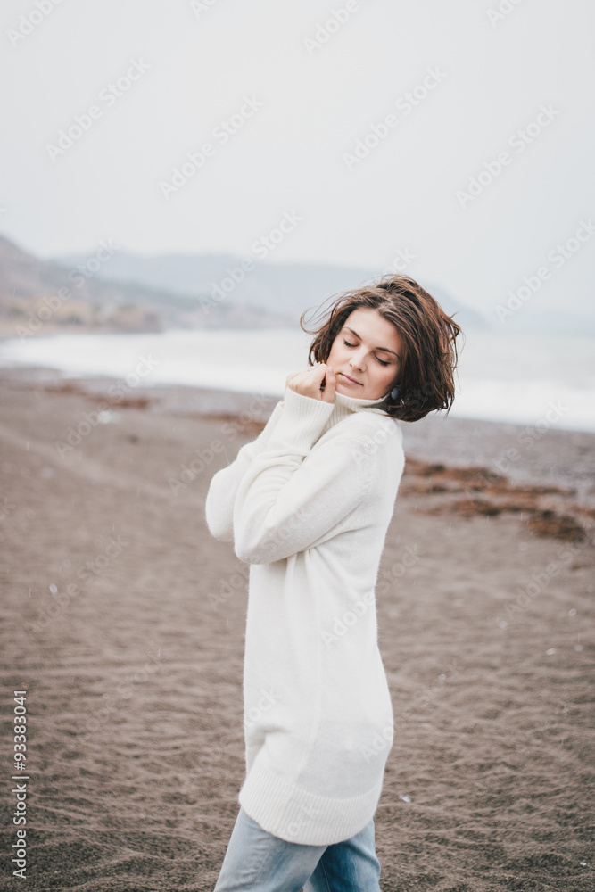 Beautiful young woman wearing white sweater and blue jeans walking on a lonely beach in a cold windy weather Beautiful young woman wearing white sweater and blue jeans walking on a lonely beach in a cold windy weather