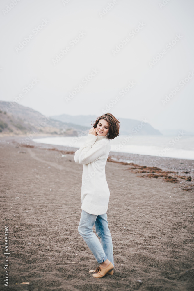 Beautiful young woman wearing white sweater and blue jeans walking on a lonely beach in a cold windy weather