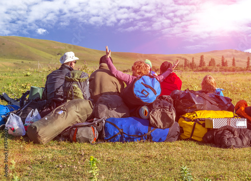 Jolly Company of Tourists Sitting on Large Heap of Travel Luggage on Green Meadow of Wild Kyrgyzstan Steppe Expressing Joy and Amazement Shining Sun above Hills on Blue Sky