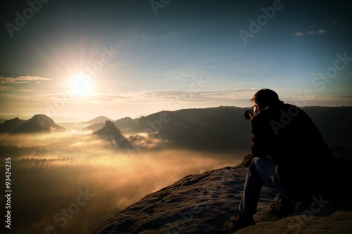 Happy tourist with camera in hands sit on peak of sandstone rock and watching into colorful mist and fog in morning valley.