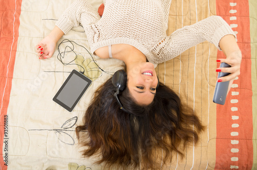 Pretty brunette wearing denim jeans white top lying down on bedsheets daydreaming with smartphone and tablet