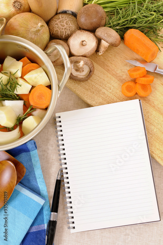 Casserole pot or stockpot crock with vegetables and herbs on kitchen worktop with blank recipe book or cookbook making preparing vegetable soup or stew photo vertical