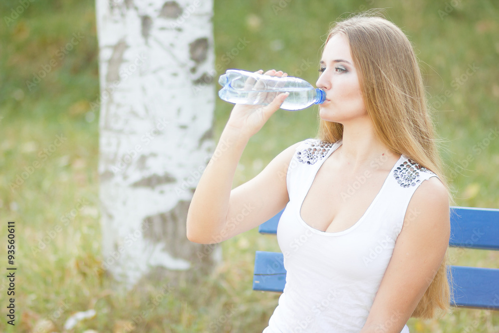 Selective focus on pretty young blond-haired woman wearing white T-shirt standing aside drinking water and smiling satisfied with her life. Park on background Selective focus on pretty young blond-haired woman wearing white T-shirt standing aside drinking water and smiling satisfied with her life. Park on background