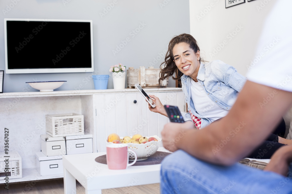 Happy couple on bed sofa in living room. Mens hand holding tv remote control while woman is playing on her tablet Happy couple on bed sofa in living room. Mens hand holding tv remote control while woman is playing on her tablet