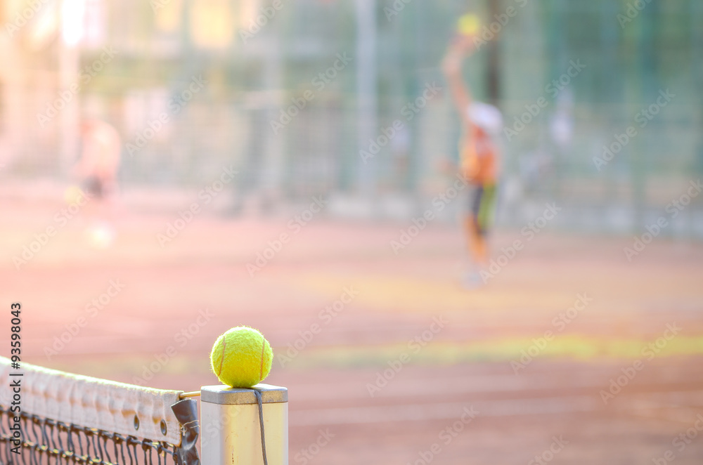 Tennis players on a clay court with a yellow ball in focus on a net post suggesting a tough tennis game with soft filter effects applied