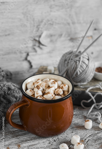 hot chocolate with marshmallows in a ceramic cup, old book, yarn and needles on a light wooden surface