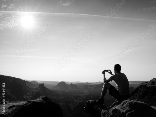 Tourist in grey t-shirt takes photos with smart phone on peak of rock. Dreamy hilly landscape below, orange pink misty sunrise in a beautiful valley below rocky mountains.