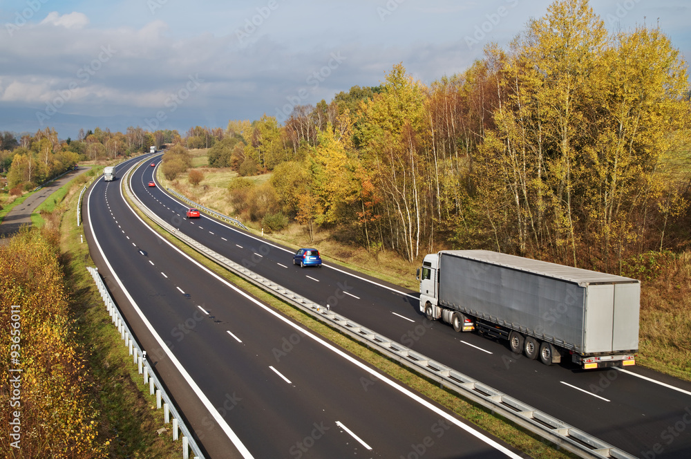 Asphalt highway with white trucks and red passenger cars in the colorful autumn landscape. Sunny day with gloomy clouds in the background. View from above.