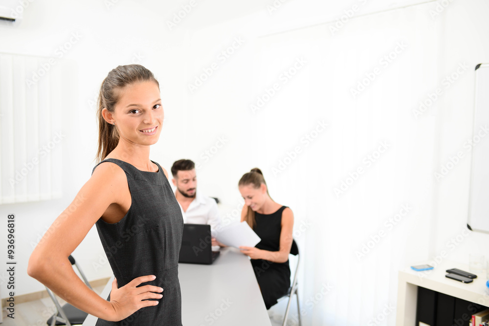 portrait of an attractive young business woman standing in office with working partner in background portrait of an attractive young business woman standing in office with working partner in background