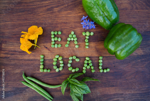 “Eat Local” phrase made of green peas on wooden background. Edible flowers, peppers, basil and green beans on a side. Organic vegetable produce at a farm. photo