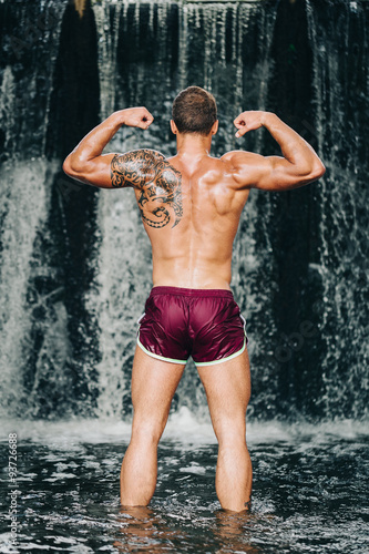 Muscular young man lifting dumbbell on waterfall