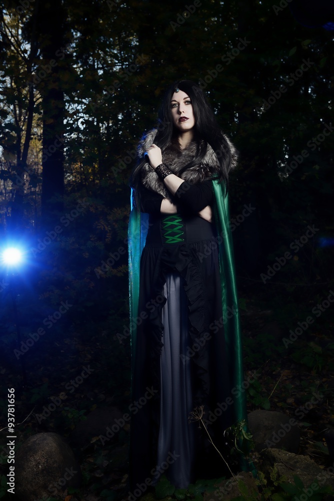 Young beautiful girl in green raincoat, looks as witch on Halloween in dark forest