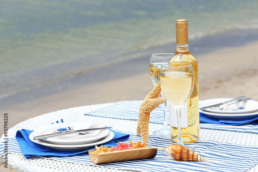Obraz premium Romantic beachside dining experience with white wine, starfish, and seashells on a striped tablecloth by the ocean
