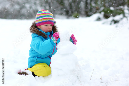 Cute toddler girl in a knitted hat and warm snowsuit playing with a snow. Little kid play having fun outdoors building snowman in the forest.
