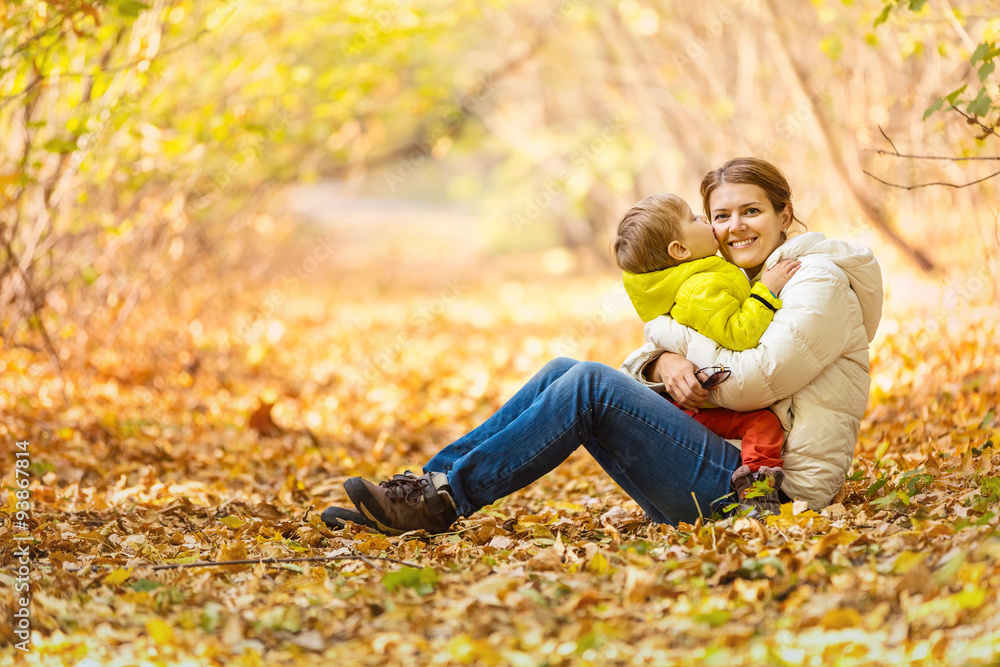 Happy woman and her little son having fun in an autumn park. The boy kissing mother while sitting on her lap. Happy woman and her little son having fun in an autumn park. The boy kissing mother while sitting on her lap.