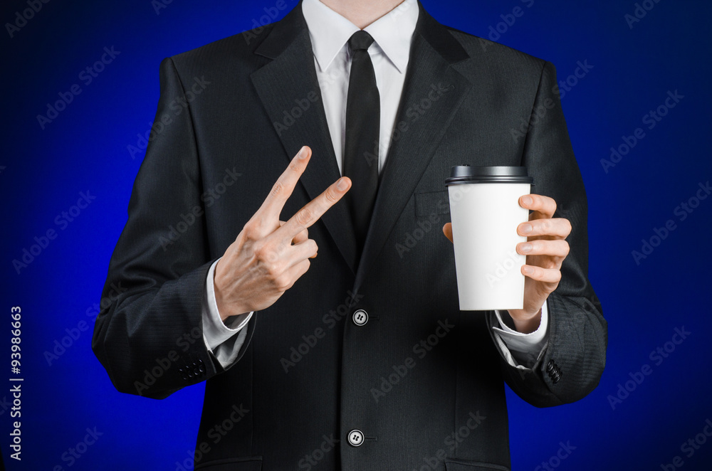 Business lunch and coffee theme: businessman in a black suit holding a white blank paper cup of coffee with a brown plastic cap on a dark blue background isolated in the studio, advertising coffee Business lunch and coffee theme: businessman in a black suit holding a white blank paper cup of coffee with a brown plastic cap on a dark blue background isolated in the studio, advertising coffee