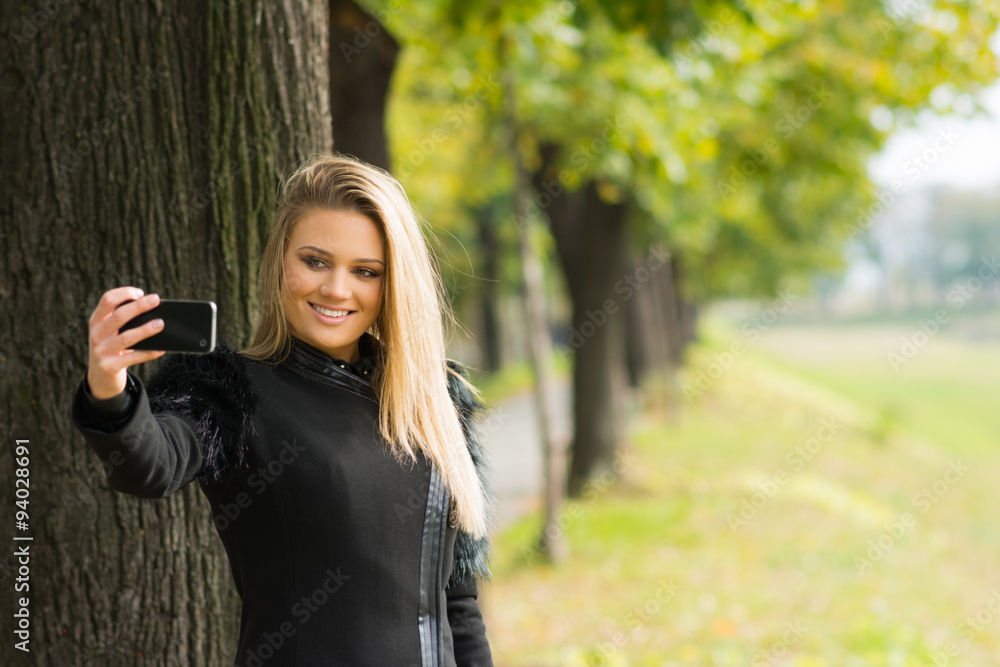 Fashionable teenage girl in black coat taking a selfie in park