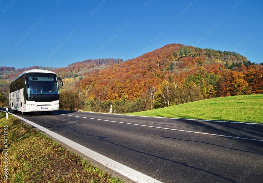 White Bus arriving at the asphalt road through the valley below the wooded mountain of glowing autumn colors. Clear sunny day with blue skies.