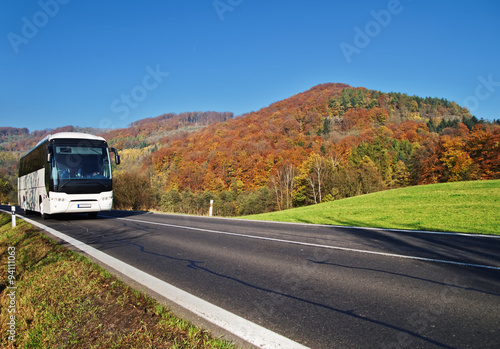 Obraz na płótnie White Bus arriving at the asphalt road through the valley below the wooded mountain of glowing autumn colors. Clear sunny day with blue skies. w salonie