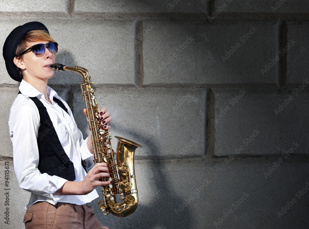 Attractive woman in hat plays saxophone on brick wall background